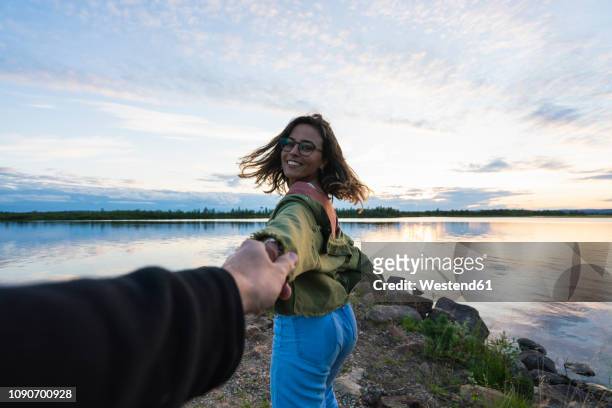 finland, lapland, happy young woman holding man's hand at the lakeside at twilight - finnland fotografías e imágenes de stock