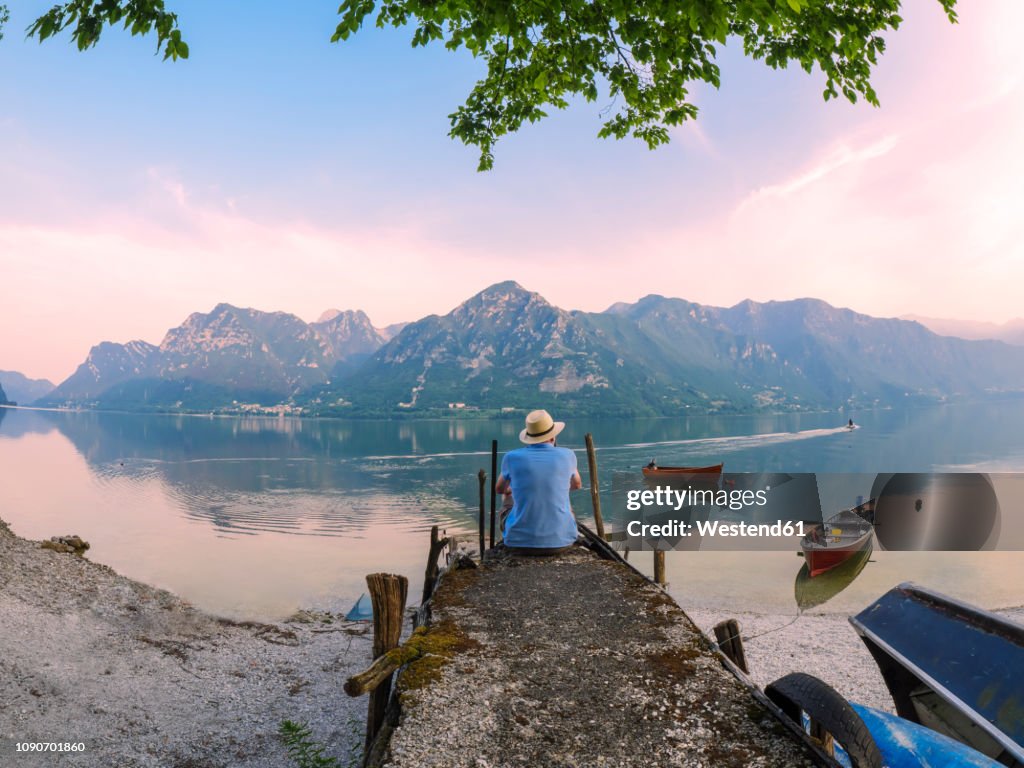 Italy, Lombardy, back view of man sitting on jetty at Lake Idro at morning twilight