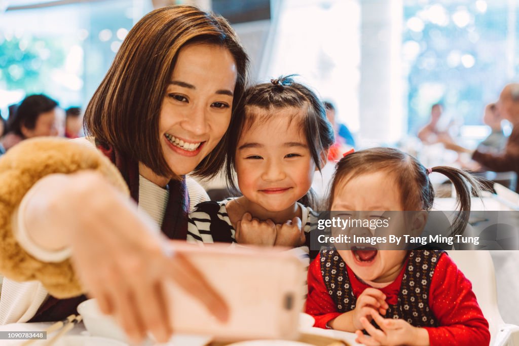 Pretty young mom taking selfies with daughters joyfully in a Chinese restaurant.