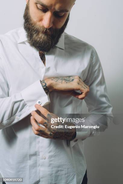 bearded young man wearing white shirt, doing up cuff links, tattoos on hands - cufflinks stock-fotos und bilder
