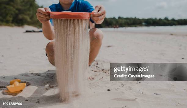 low section of boy sieving sand while crouching at beach - passoire photos et images de collection