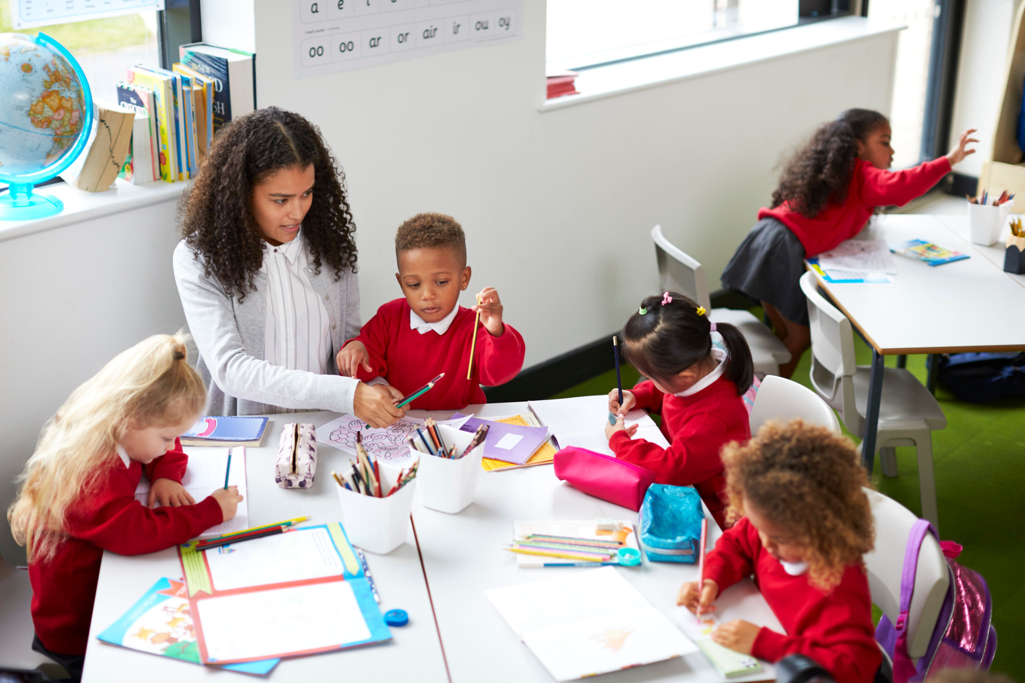 Elevated view of female kindergarten teacher sitting at a table helping four children during a lesson Elevated view of female kindergarten teacher sitting at a table helping four children during a lesson