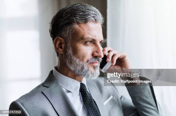 a portrait of a mature attractive businessman with smartphone standing in an office, making a phone call. - grey jacket stock pictures, royalty-free photos & images