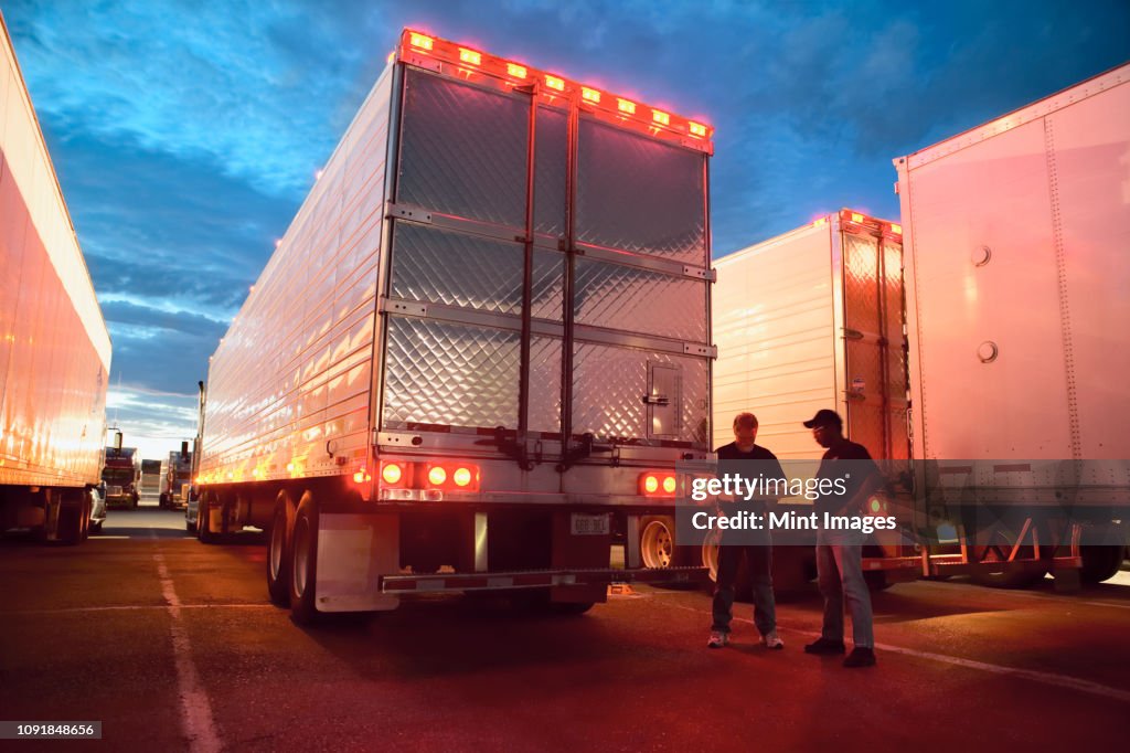Two truck drives checking dispatch papers while standing next to truck trailers in a large parking lot at night.