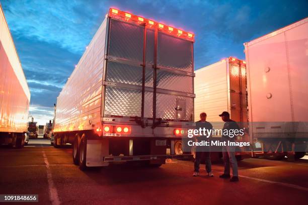 two truck drives checking dispatch papers while standing next to truck trailers in a large parking lot at night. - truck driver stock pictures, royalty-free photos & images