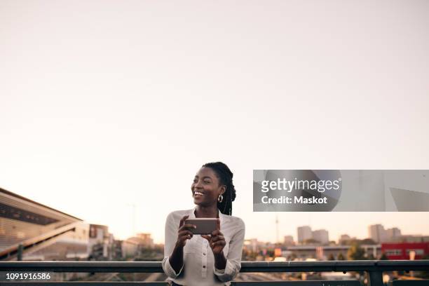 smiling young woman looking away while holding mobile phone against clear sky in city - clear sky stock-fotos und bilder