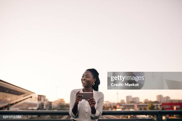 smiling young woman looking away while holding mobile phone against clear sky in city - mobile technology imagens e fotografias de stock