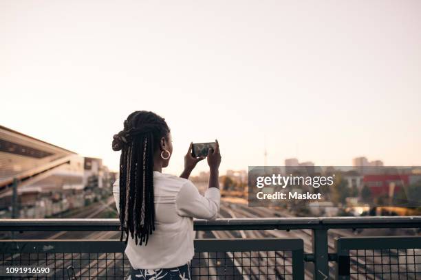 young woman photographing through mobile phone while standing on bridge against clear sky - frau zopf hinten stock-fotos und bilder