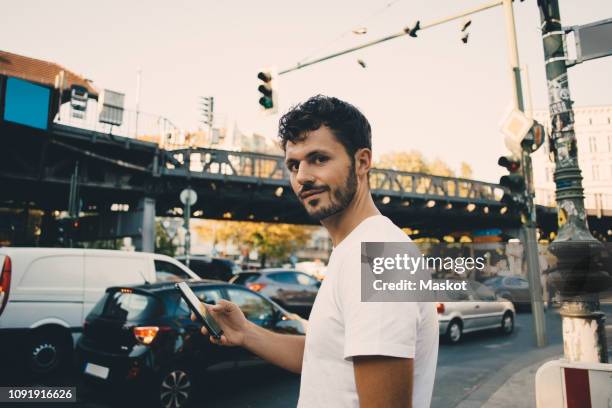 portrait of young man holding mobile phone while standing on sidewalk in city - cool cars stockfoto's en -beelden