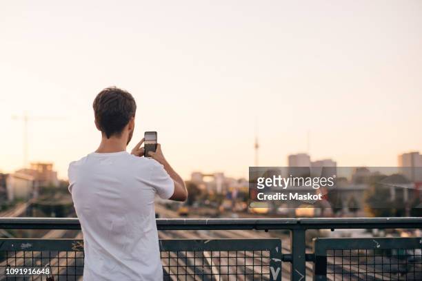 rear view of young man photographing city through mobile phone while standing on bridge against clear sky - rückansicht stock-fotos und bilder