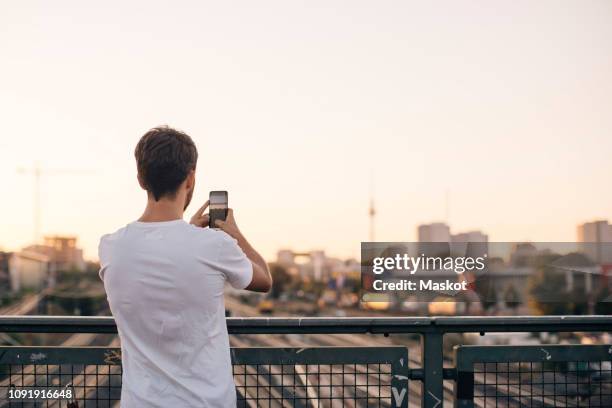 rear view of young man photographing city through mobile phone while standing on bridge against clear sky - back foto e immagini stock