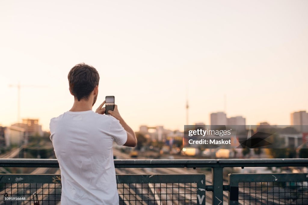 Rear view of young man photographing city through mobile phone while standing on bridge against clear sky