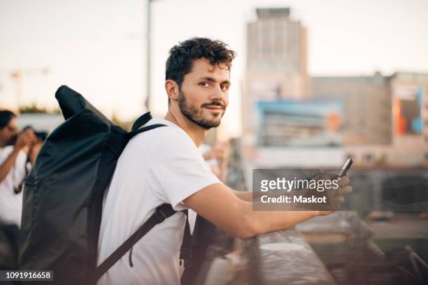 side view portrait of young man holding mobile phone while leaning on railing at bridge - leben in der stadt stock-fotos und bilder