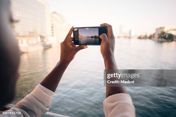 cropped image of young woman photographing river through mobile phone in city - black hand holding phone stock-fotos und bilder