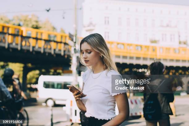 young woman using mobile phone while walking by street in city - berlin stock-fotos und bilder