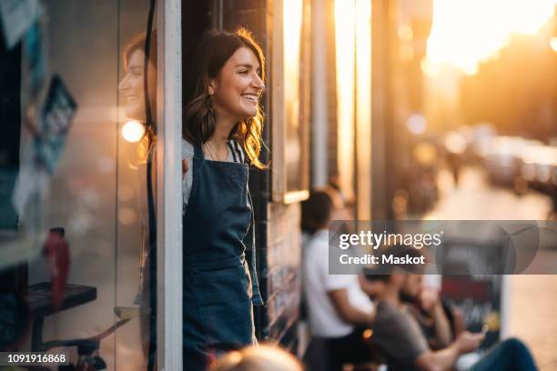 smiling female owner looking away while standing at entrance of deli - outdoor cafe stock-fotos und bilder