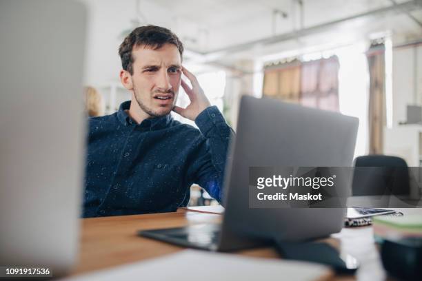 confused businessman looking at laptop while sitting at desk in office - frustration stock-fotos und bilder