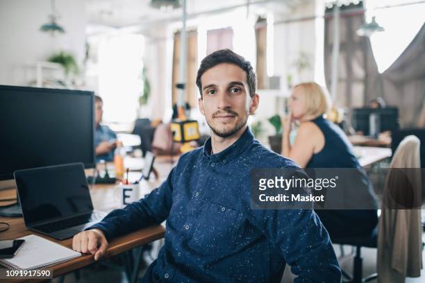 portrait of confident young businessman sitting at desk in creative office - personas en el fondo fotografías e imágenes de stock