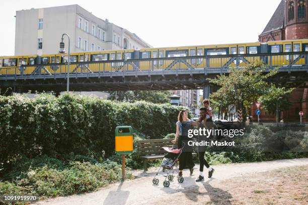 young man carrying daughter on shoulders while woman pushing baby stroller at footpath against railway bridge - berlin stadt stock-fotos und bilder