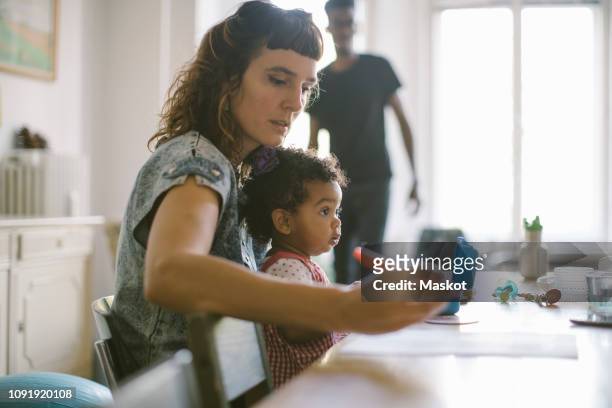 woman working while sitting with daughter at table in house - madre capofamiglia foto e immagini stock