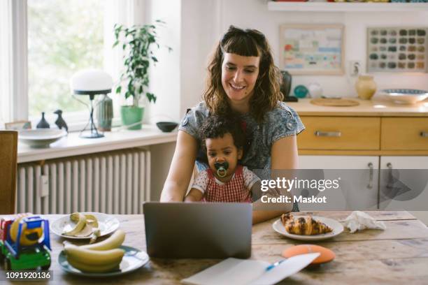 smiling woman using laptop while sitting with daughter at dining table in house - busy toddlers stock pictures, royalty-free photos & images