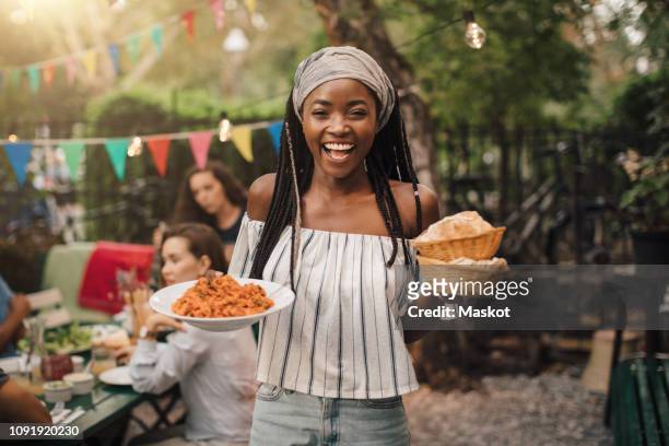 portrait of smiling young woman carrying food while standing in backyard during garden party - dinner party stock pictures, royalty-free photos & images