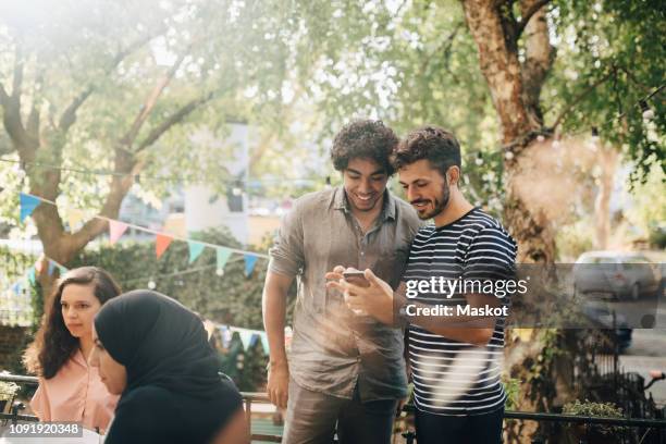 smiling young man showing mobile phone to male friend while standing in balcony during party - man showing phone stock pictures, royalty-free photos & images