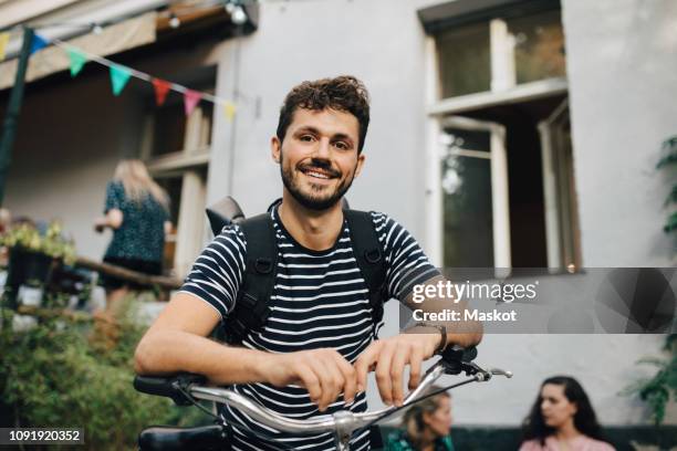 portrait of smiling young man leaning on bicycle in backyard - adulto joven fotografías e imágenes de stock