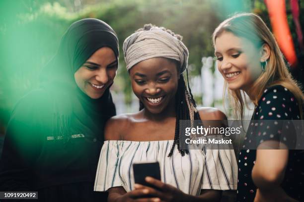 smiling multi-ethnic female friends looking at mobile phone while standing in backyard - religion stock-fotos und bilder