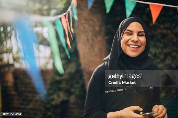 portrait of smiling young woman in hijab holding mobile phone at backyard - arab home stock pictures, royalty-free photos & images