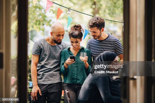 smiling young woman showing mobile phone to male friends while standing in balcony during party - male cellphone stock-fotos und bilder