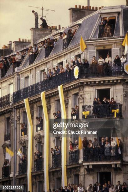 View of onlookers crowded onto balconies of an unidentified apartment building, decorated with white and yellow bunting for Pope John Paul II's...