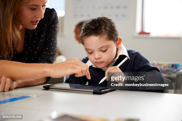 年輕的女教師與唐氏綜合症小學生坐在辦公桌前使用平板電腦在小學教室, 前面的視野, 關閉
