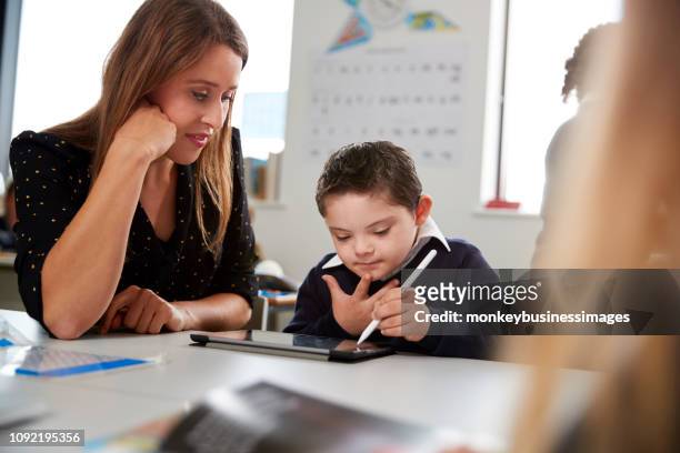 Young female teacher working with a Down syndrome schoolboy sitting at desk in a primary school classroom, selective focus