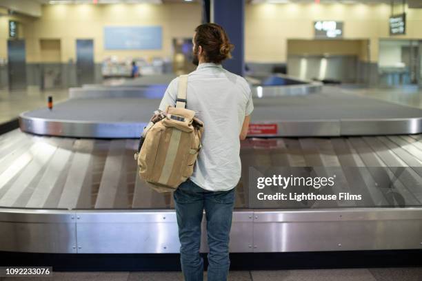 hip young man stands alone waiting for luggage by empty baggage carousel. - baggage claim stock pictures, royalty-free photos & images