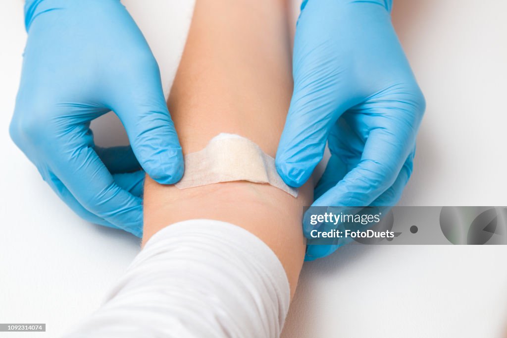 Doctor in blue rubber protective gloves putting an adhesive bandage on young woman's arm vein after blood test or injection of vaccine. First aid. Medical, pharmacy and healthcare concept. Closeup.