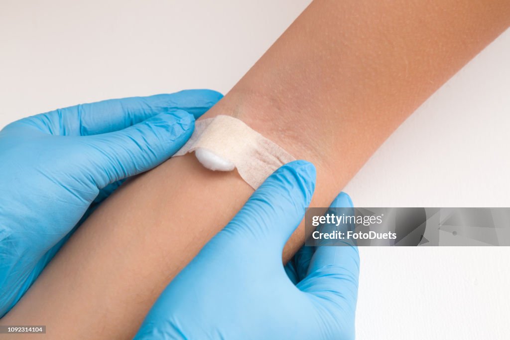 Doctor in blue rubber protective gloves putting an adhesive bandage on young woman's arm vein after blood test or injection of vaccine. First aid. Medical, pharmacy and healthcare concept. Closeup.