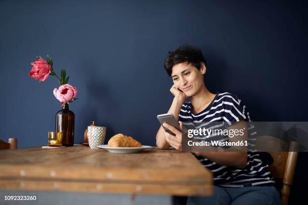 smiling woman at home sitting at wooden table checking cell phone - blue wooden table stock-fotos und bilder