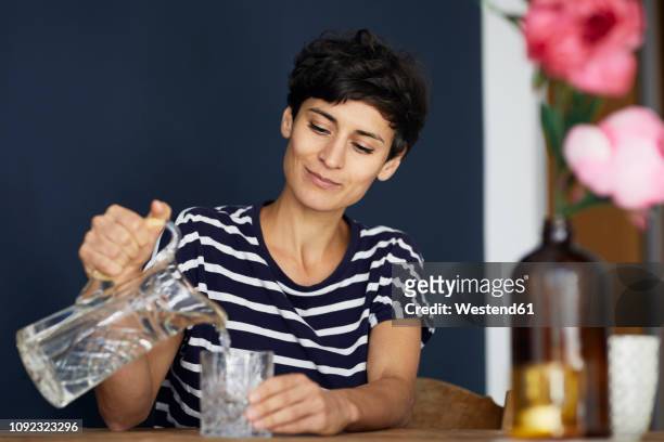 woman at home sitting at wooden table pouring water into glass - wasser stock-fotos und bilder
