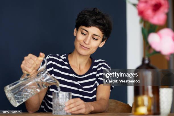 woman at home sitting at wooden table pouring water into glass - gießen wasser stock-fotos und bilder