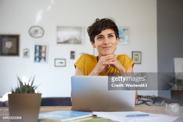 portrait of smiling woman at home sitting at table using laptop - beschaulichkeit stock-fotos und bilder