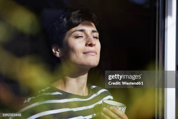 woman with cup of coffee at the window enjoying the sunshine - enjoy stockfoto's en -beelden