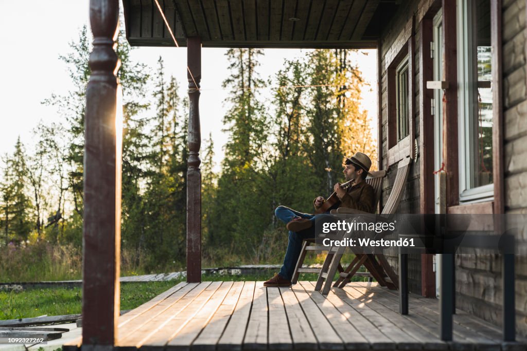 Young man sitting on veranda of a wood house, playing the ukulele