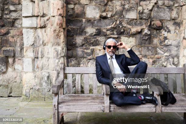 portrait of well-dressed senior businessman with sunglasses and headphones sitting on bench - excéntrico fotografías e imágenes de stock