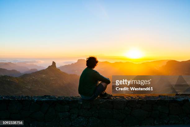 spain, canary islands, gran canaria, back view of man sitting on a wall watching sunset over mountainscape - tramonto foto e immagini stock