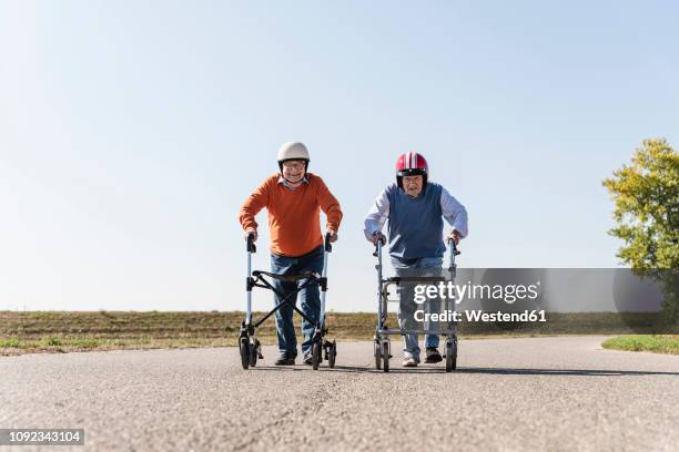 two old friends wearing safety helmets, competing in a wheeled walker race - rollator stock-fotos und bilder