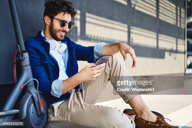 smiling young man sitting on the ground besides his electric scooter using cell phone - ventenne foto e immagini stock