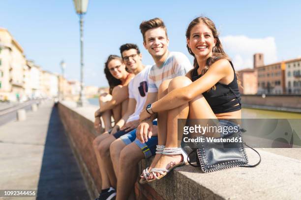 italy, pisa, group of four happy friends sitting on a wall along arno river - chico ciudad fotografías e imágenes de stock