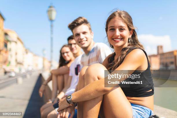 italy, pisa, group of four happy friends sitting on a wall along arno river - boy river looking at camera stock pictures, royalty-free photos & images