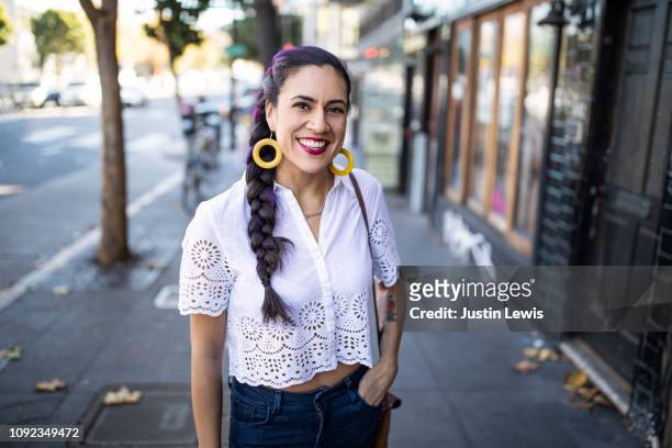millennial latina stands alone on city sidewalk, smiling and looking at camera, wearing white lace blouse and bright yellow hoop earrings - woman 30 outside stock-fotos und bilder